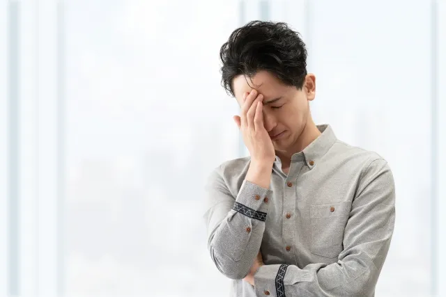 立ちながら右手で顔を覆って悩んでいるグレーのYシャツの男性