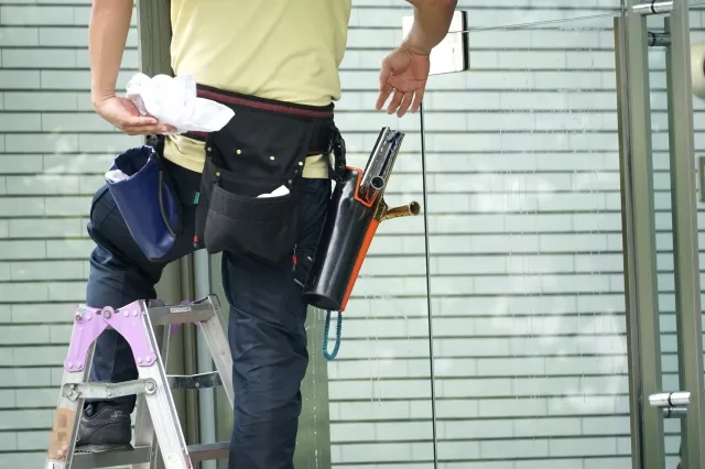 腰に付けている道具袋に窓清掃用の道具を入れて脚立に上って大きな窓を清掃しようとしている男性