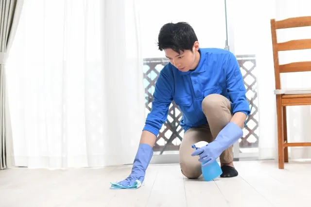 青い長そでの作業着を着た男性がゴム手袋をして床の清掃をしている